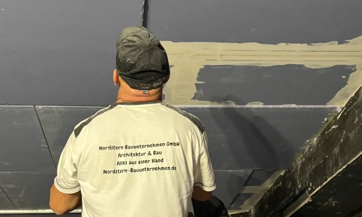 Handwerker im T-Shirt mit Nordstern-Aufdruck, von hinten gesehen, unter einer Trockenbaudecke mit verspachtelten Fugen