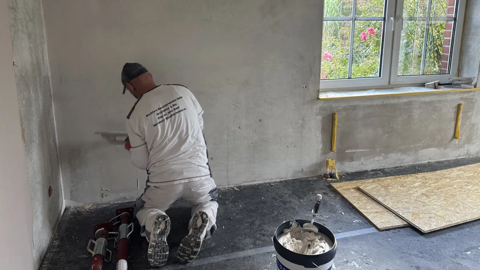 Handwerker in Nordstern-Arbeitskleidung kniet vor einer Betonwand und trägt mit der Glättkelle Gipsputz auf, daneben ein Eimer mit Putzmaterial
